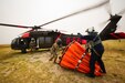 Two men, one military and one a civilian, load a water bucket onto a helicopter.