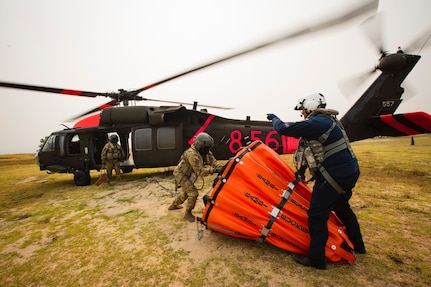 Two men, one military and one a civilian, load a water bucket onto a helicopter.