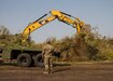 A construction truck loads tree debris onto a Utah National Guard flatbed truck to be hauled away to a local landfill while a Utah National Guard soldier looks on.
