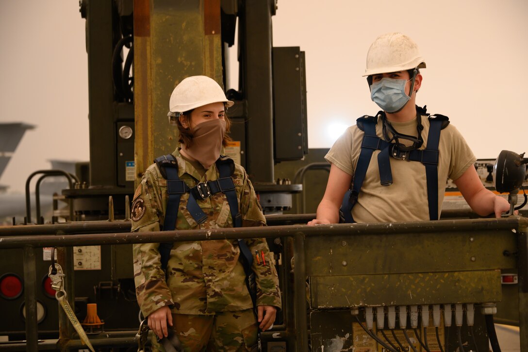 Two airmen stand on a large lift condor with a yellowish, smokey sky.