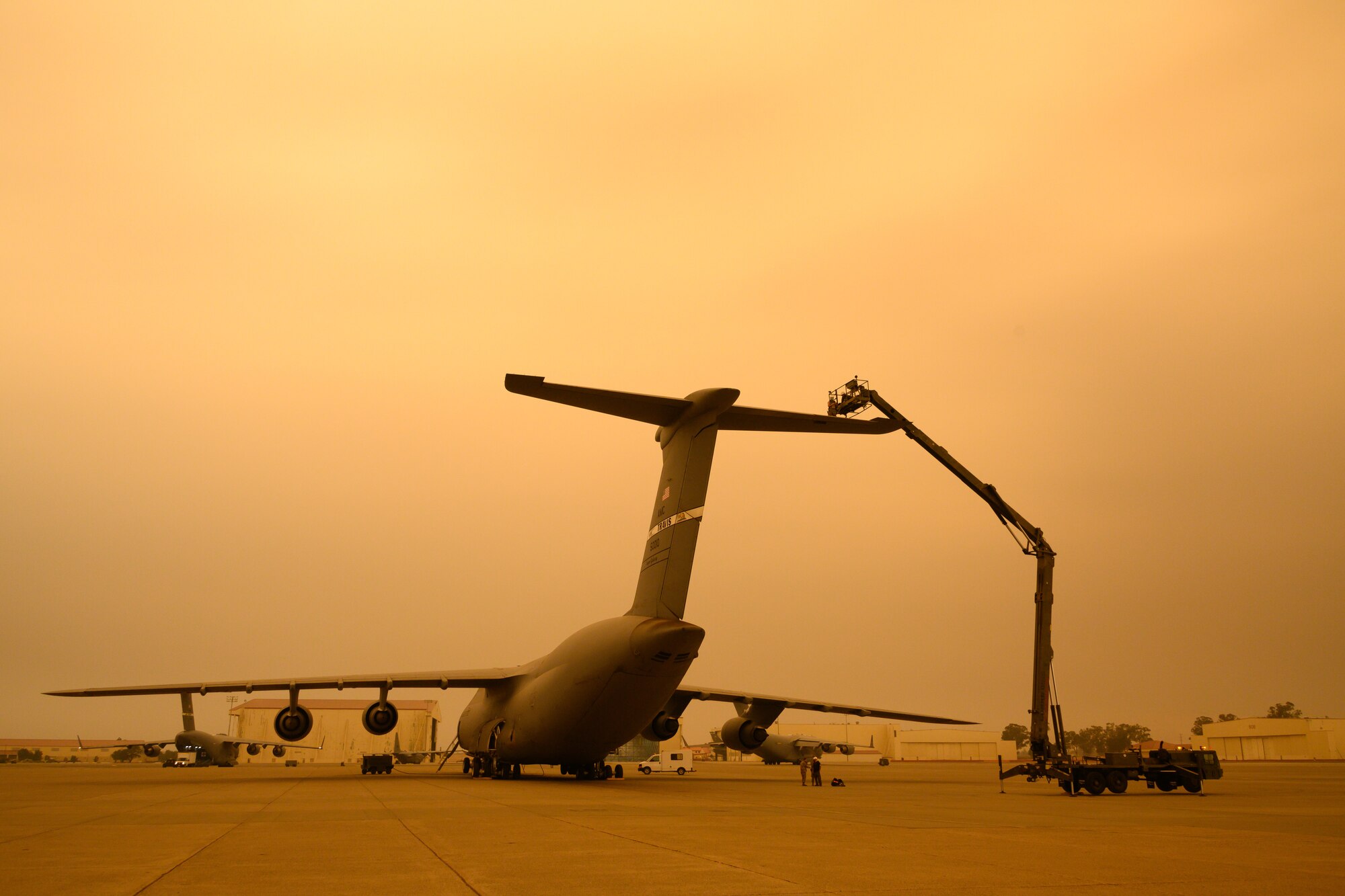 Airmen are lifted in a large condor lift above a C-5M tail with a smokey, yellowish sky.