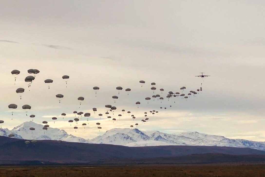 A large group of soldiers free fall wearing parachutes with snowy mountains in the background.