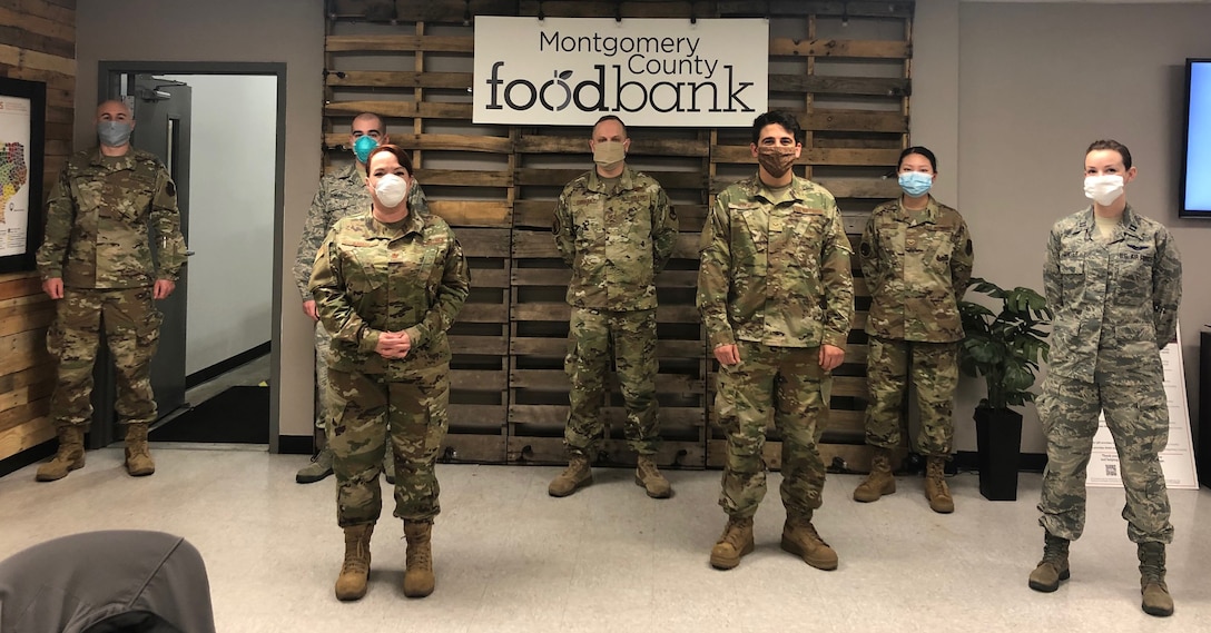 Military personnel wearing masks stand in front of a Montgomery County Food Bank sign