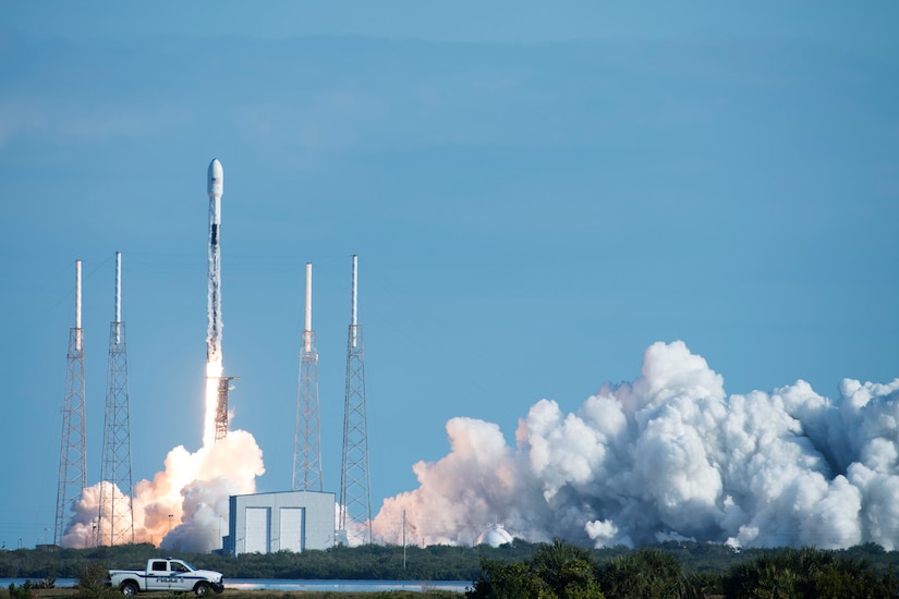 A rocket launches against a blue sky.  Flames and smoke billow out underneath.