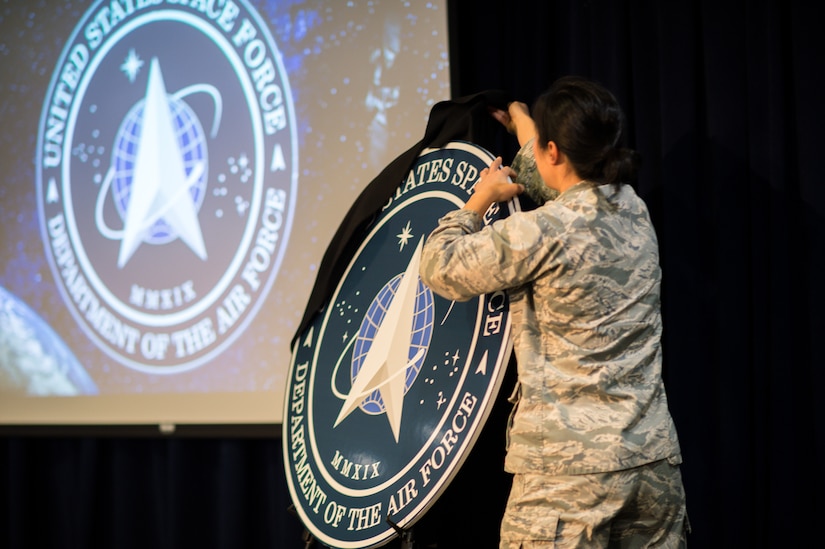 An airman pulls a black sheet off an easel that holds a circular cutout featuring the seal of the U.S. Space Force.