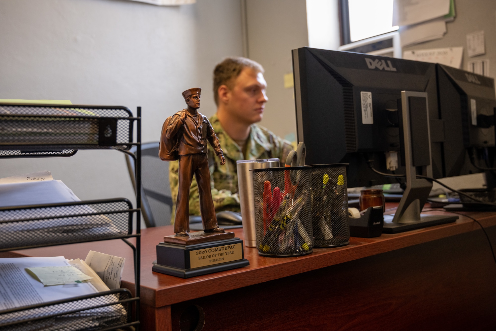 A statue that says "US Navy Sailor of the Year Finalist" sits on the corner of an office desk while a Navy service member works at their computer.