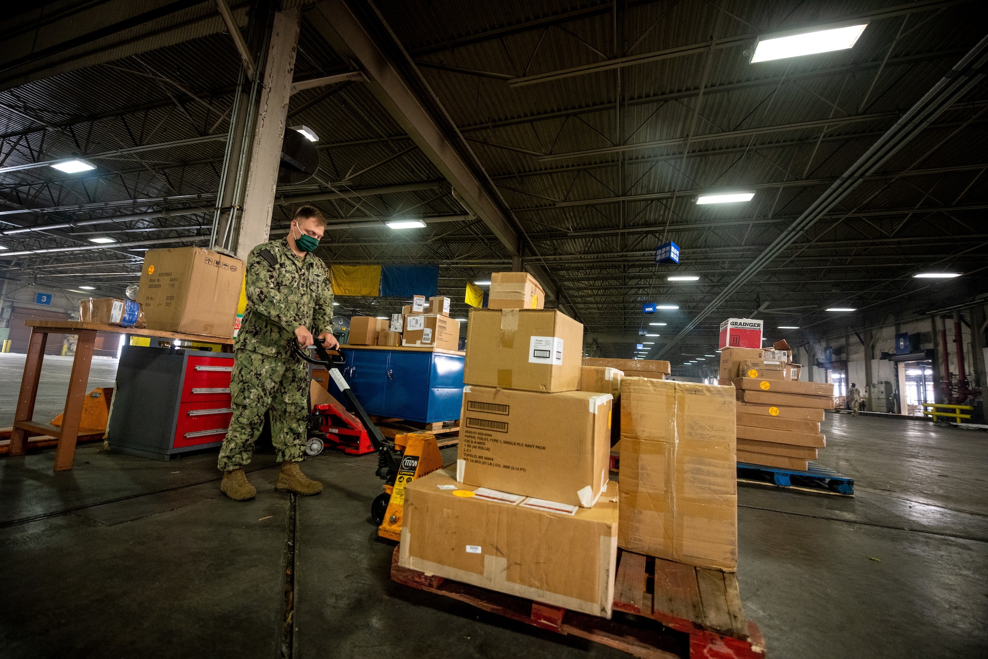 A Navy service member moves a small pallet of boxes inside a large warehouse.