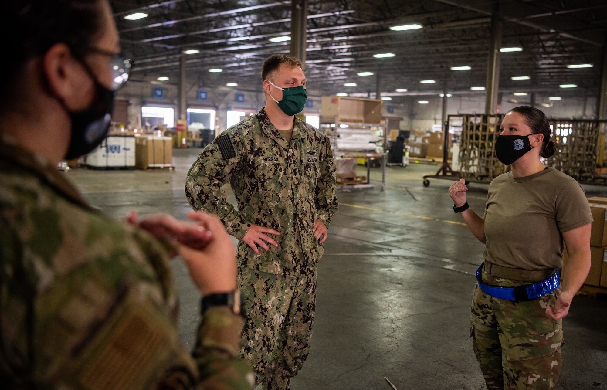 A Navy service member talks with Air Force members inside a large warehouse full of boxes.