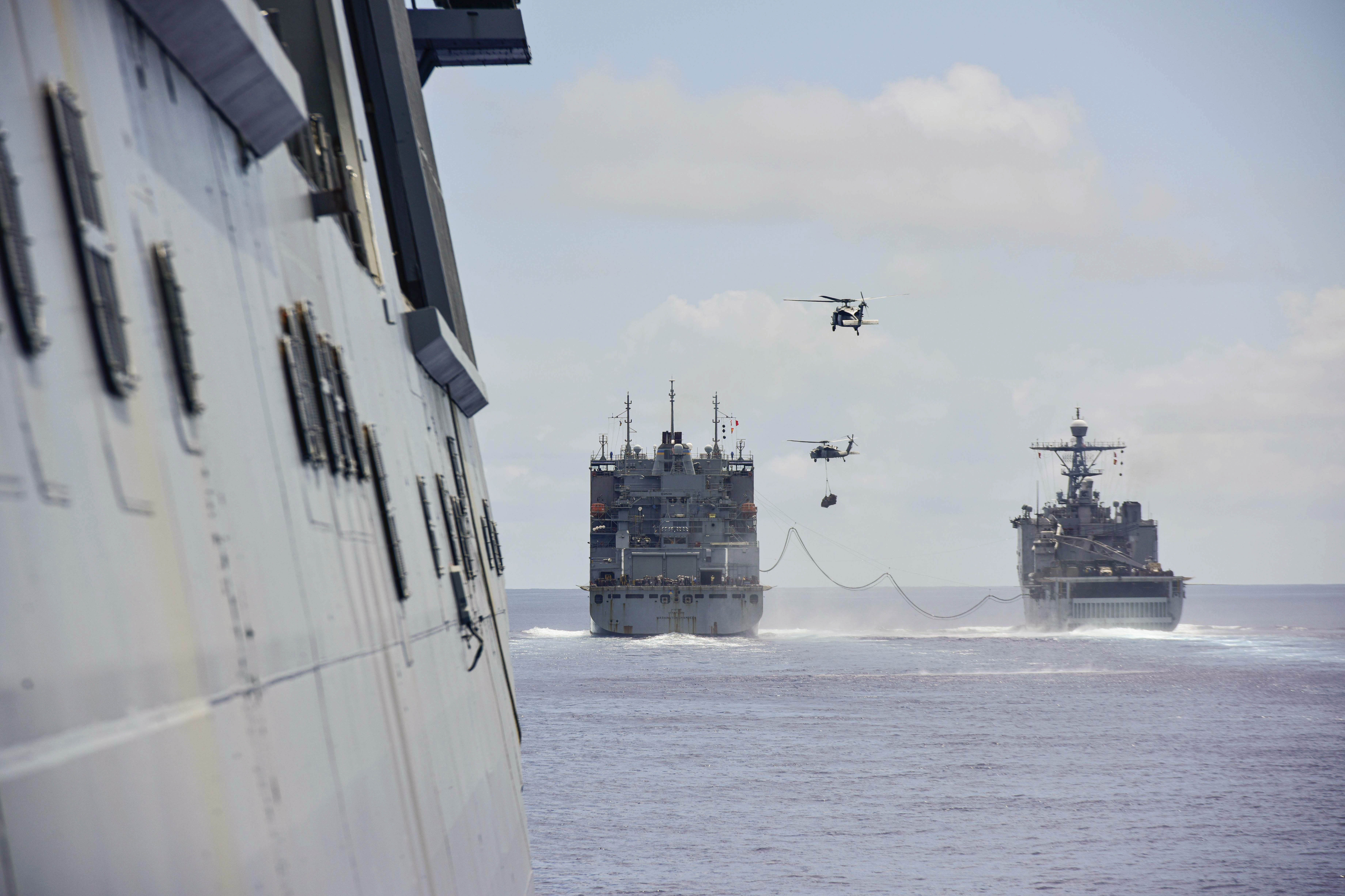 Two ships sail next to each other as two helicopters fly above.