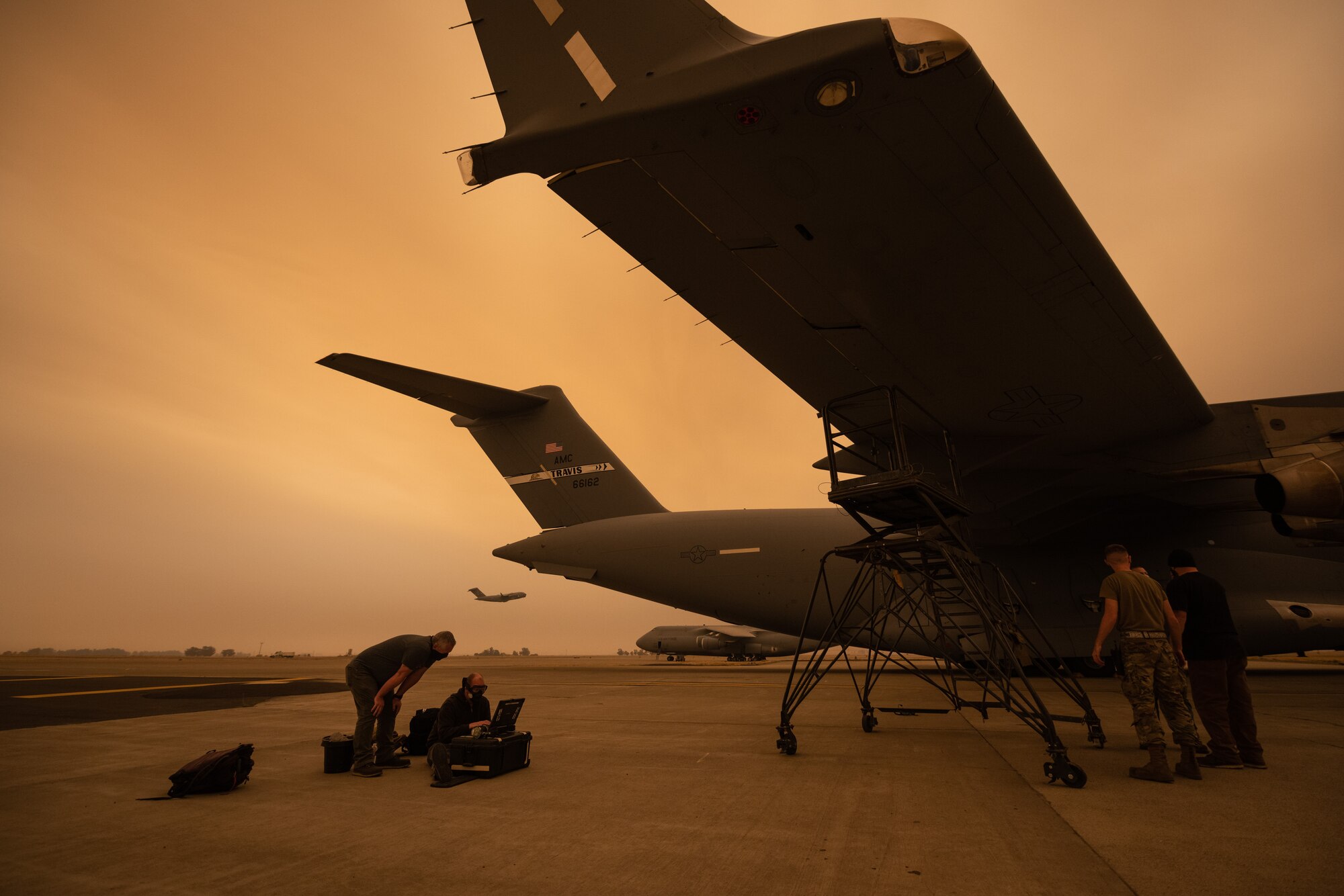 Airmen work on a wing of a C-17 while another C-17 takes off in the background.