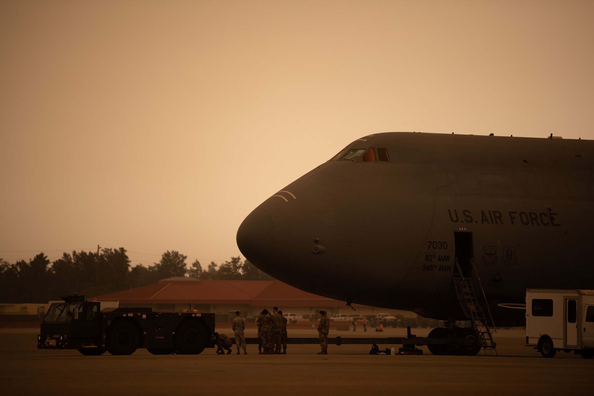 Airmen attach a towing vehicle to a C-5M on the flight line with a yellowish, smokey sky.