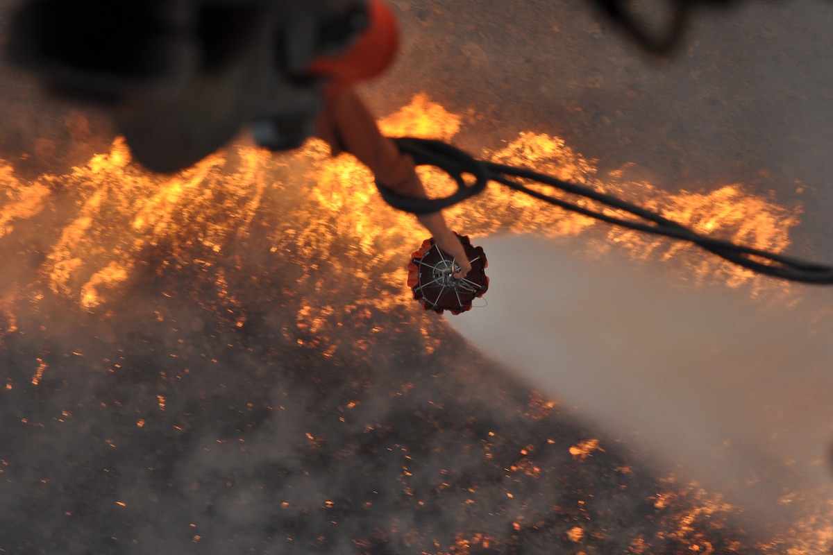 A helicopter empties  a water bucket over flames and smoke during a wildfire.