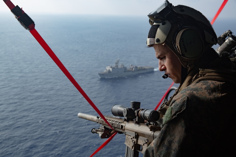 A person with a gun looks out of a helicopter over the ocean.