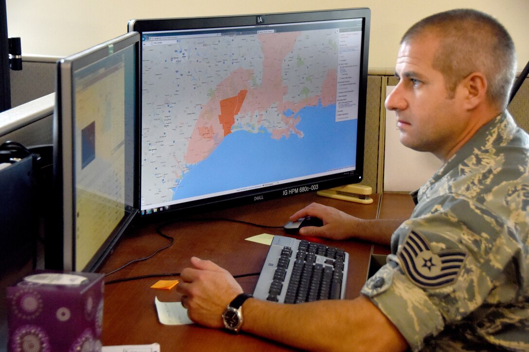 A man in a military uniform sits before two computer screens; one displays a map while the other displays related data.