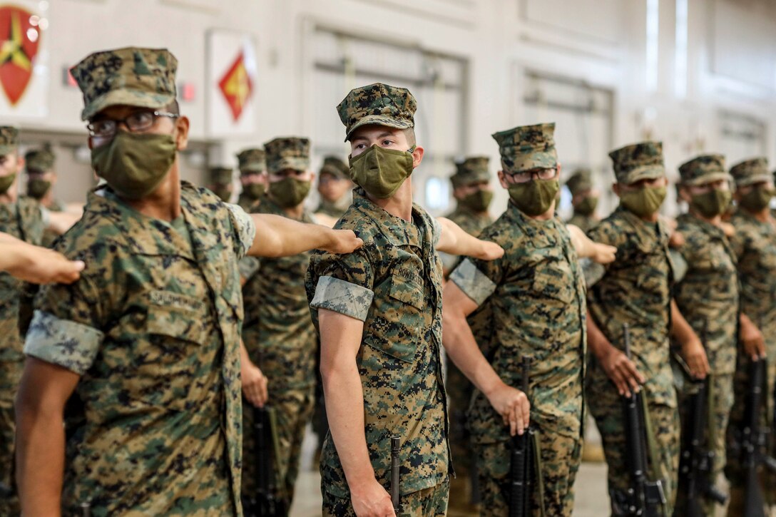 Marine recruits wearing protective gear stand in a line holding up their left arms.