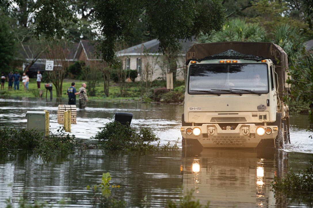 Soldiers help residents in high water as a military vehicle drives through a flooded area.