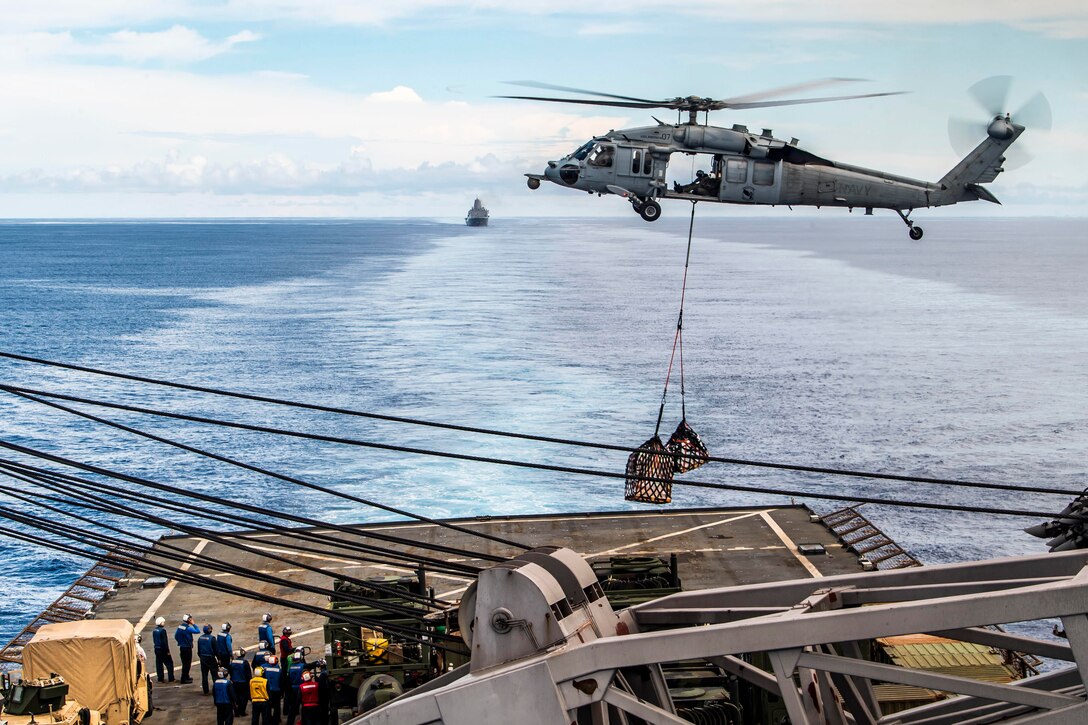 A helicopter prepares to drop cargo onto a ship’s deck.