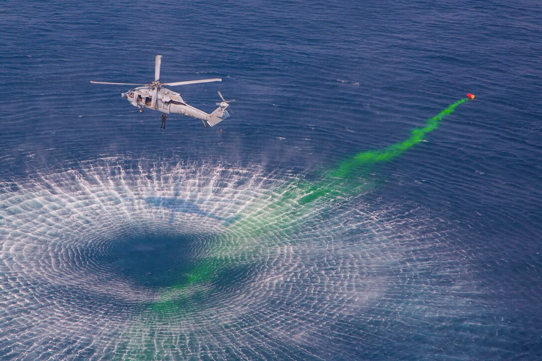 A helicopter pulls a service member from the water with green smoke from a mine passing by.