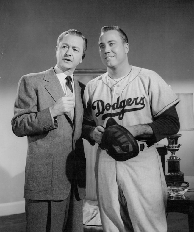 A man in a suit stands with his arm over the shoulder of a man wearing a baseball uniform.