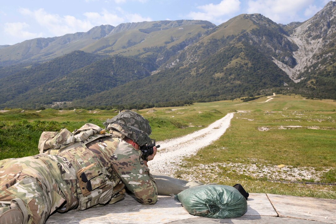 A soldier lays on the ground and fires a weapon towards mountains.