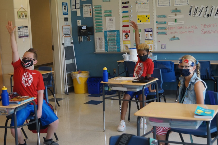 Kids wearing face masks raising their hands in a classroom.