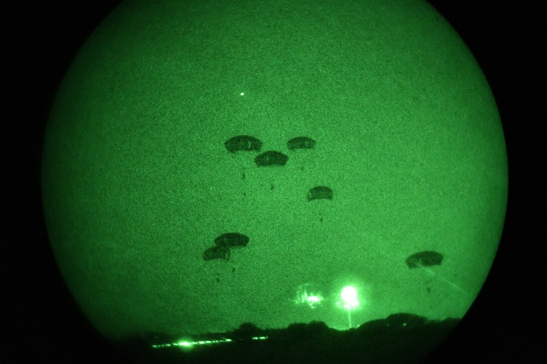 Soldiers illuminated by green light freefall wearing parachutes.