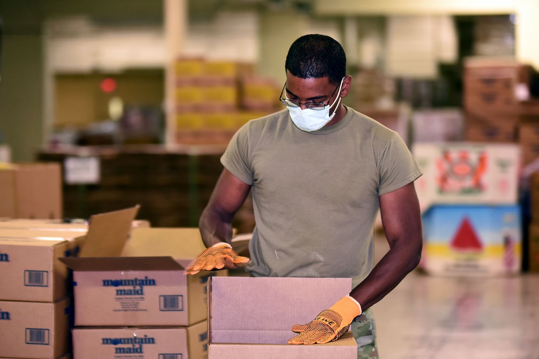 A soldier wearing a face mask and gloves prepare meal boxes for distribution at a food bank.