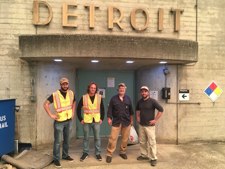 Four men stand in front of doors, with a sign above them that reads Detroit.