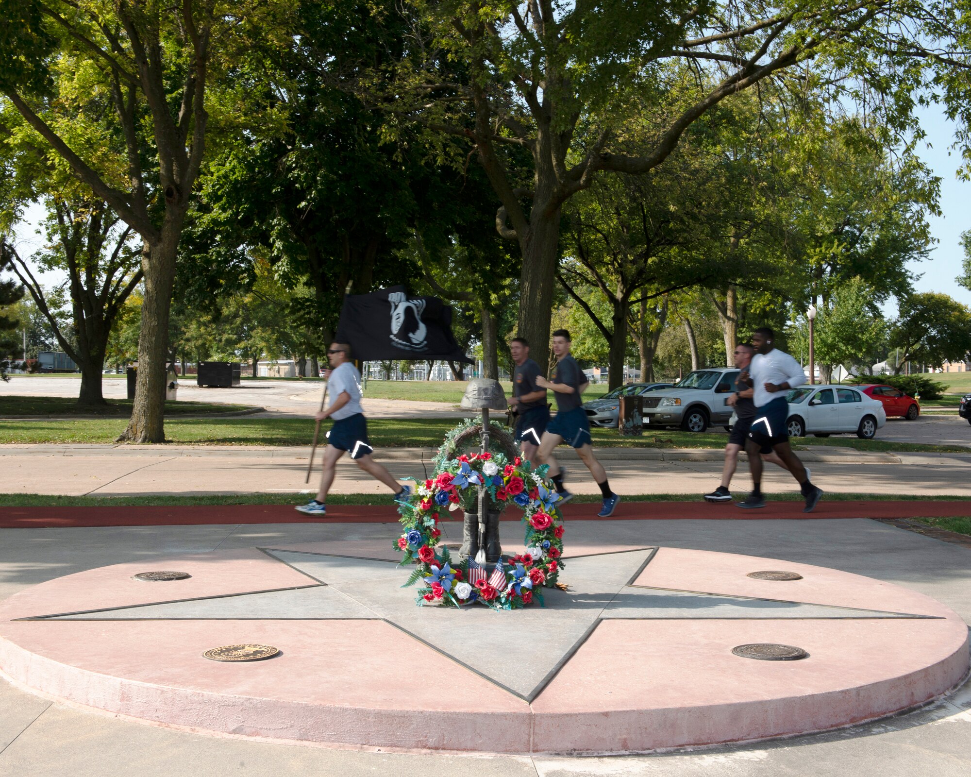 in the front of photograph is the Prisoner of War/Missing in Action Memorial with a large wreath leaning against it. In the background an Airman running while carrying the POW/MIA flag running with four other Airmen running behind him