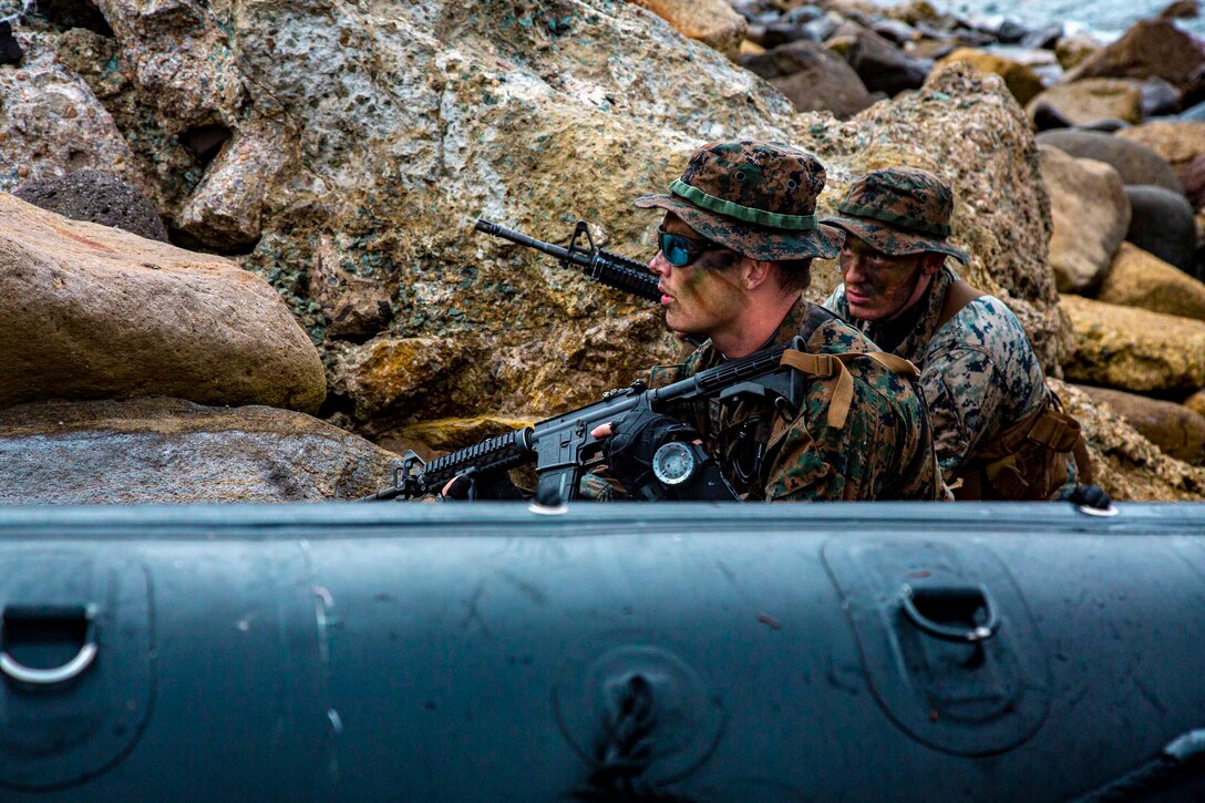 Two Marines wearing camouflage paint carry rifles while positioned against rocks.