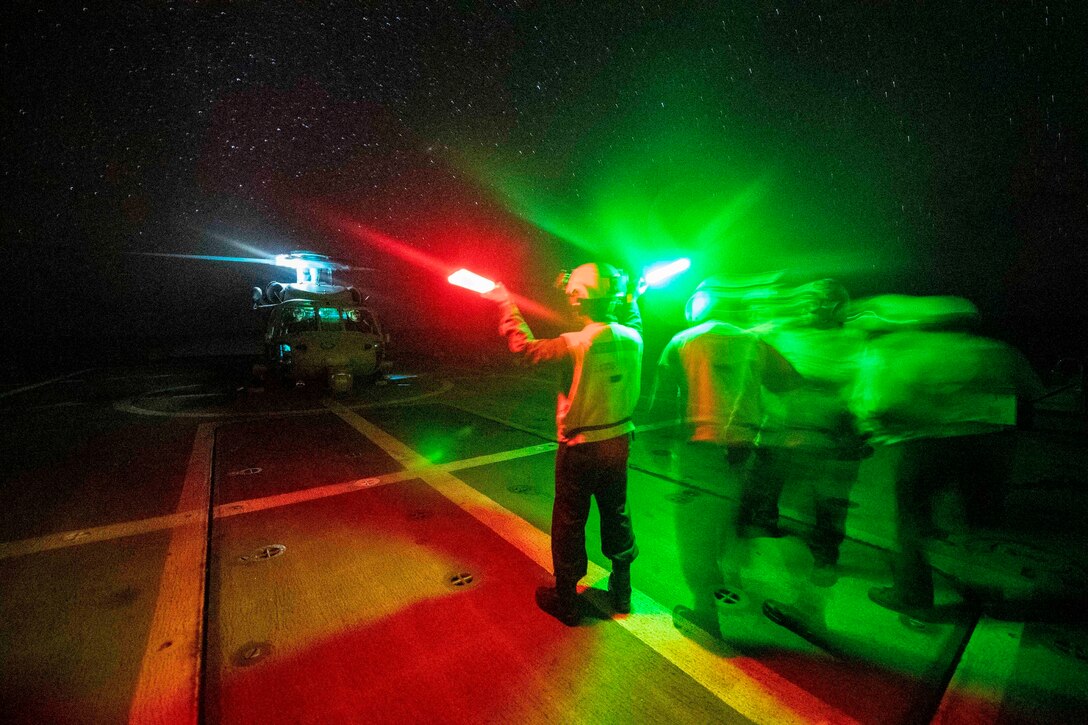 Sailors bathed in red and green colors signal a helicopter onto the deck of a ship.