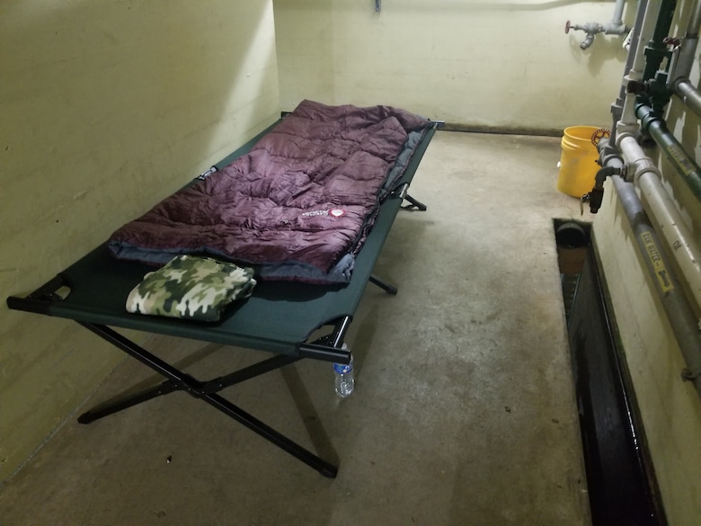 A cot with a sleeping bag sits in a concrete hallway.