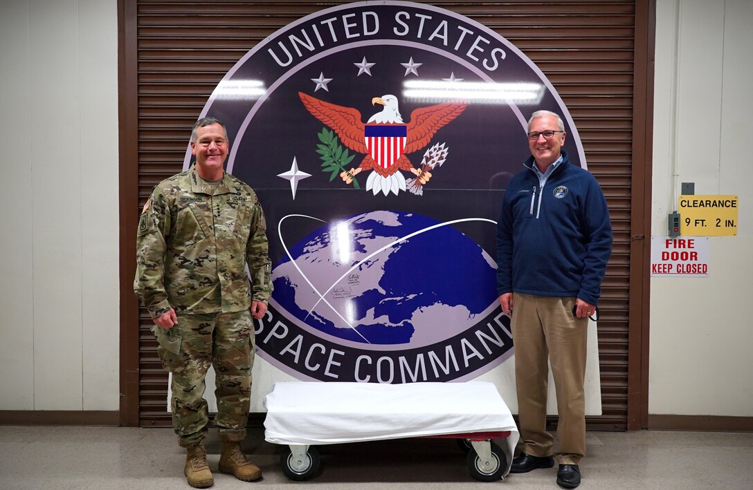 A circular U.S. Space Command seal stands nearly seven feet tall and wide between two men who smile for a photo beside it.