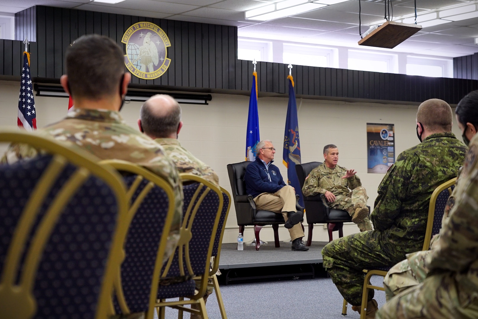 An audience of military members sit with their backs to the foreground of a photo that focuses on two men sitting on-stage in dark blue chairs.