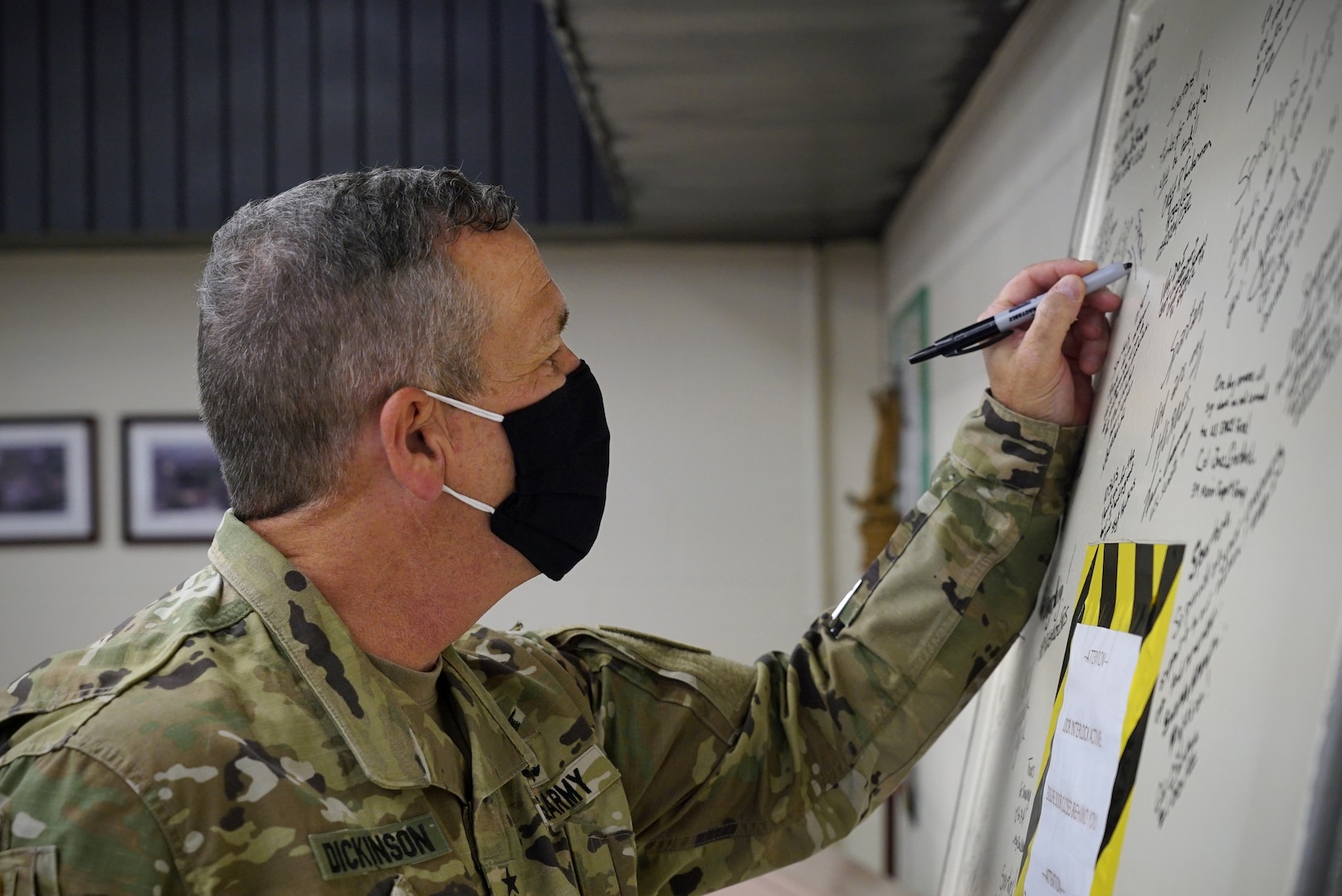 A man in military uniform uses his left hand to leave his signature in permanent marker on a door leaning up against the wall in front of him.