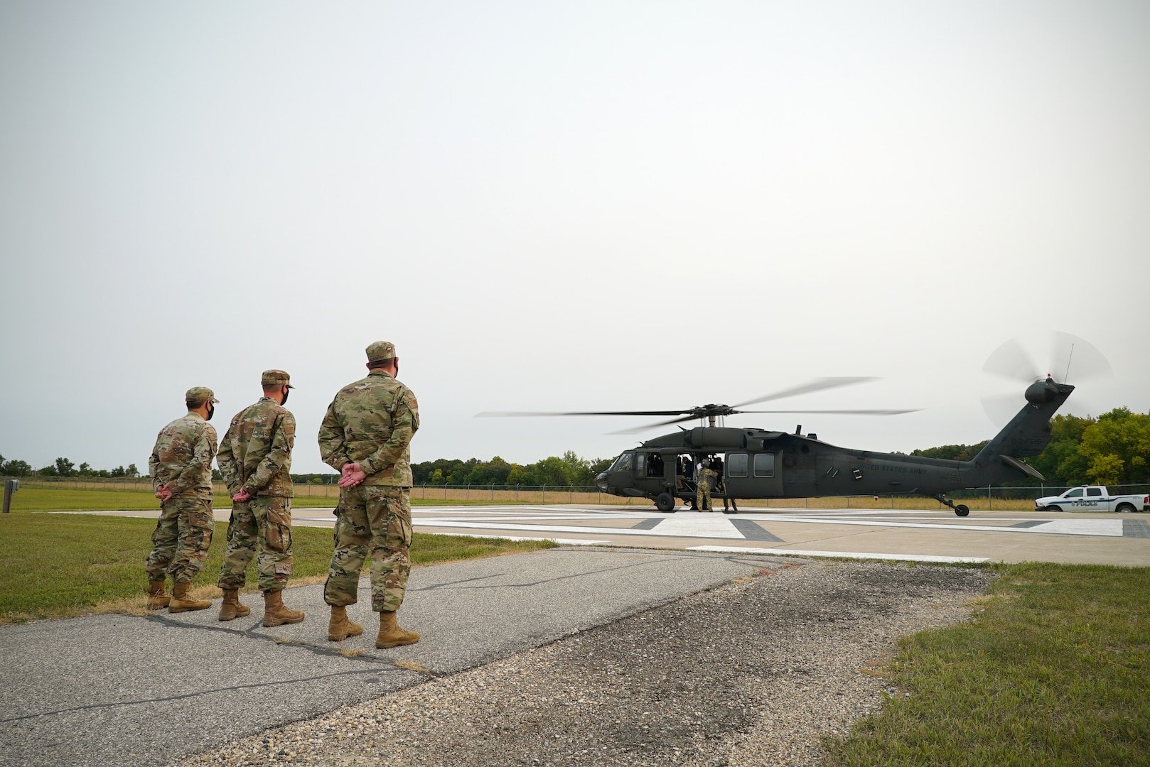 Three men in military uniform stand at parade rest on the left-hand side of the screen, awaiting the departure of a UH-60 Black Hawk seen on the right-hand of the screen.