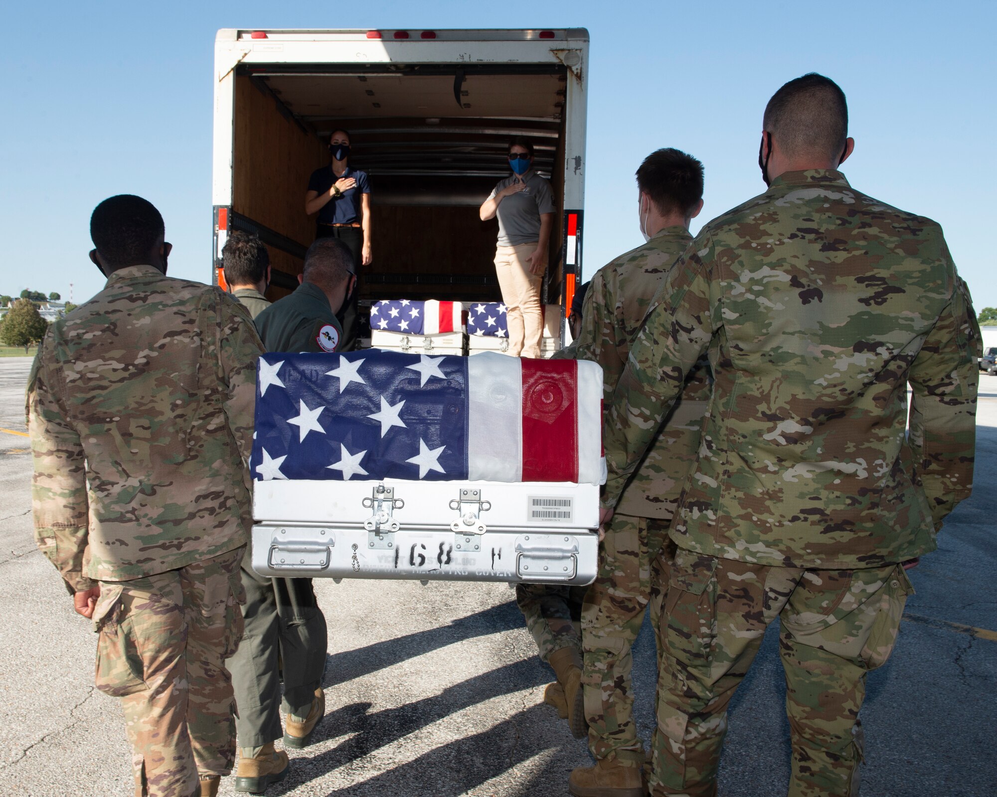 in the foreground, six Airmen carry a U.S. flag draped transfer case to a transportation truck in the background, two civilians stand inside with hands over there hearts, inside the truck are two previously loaded transfer cases