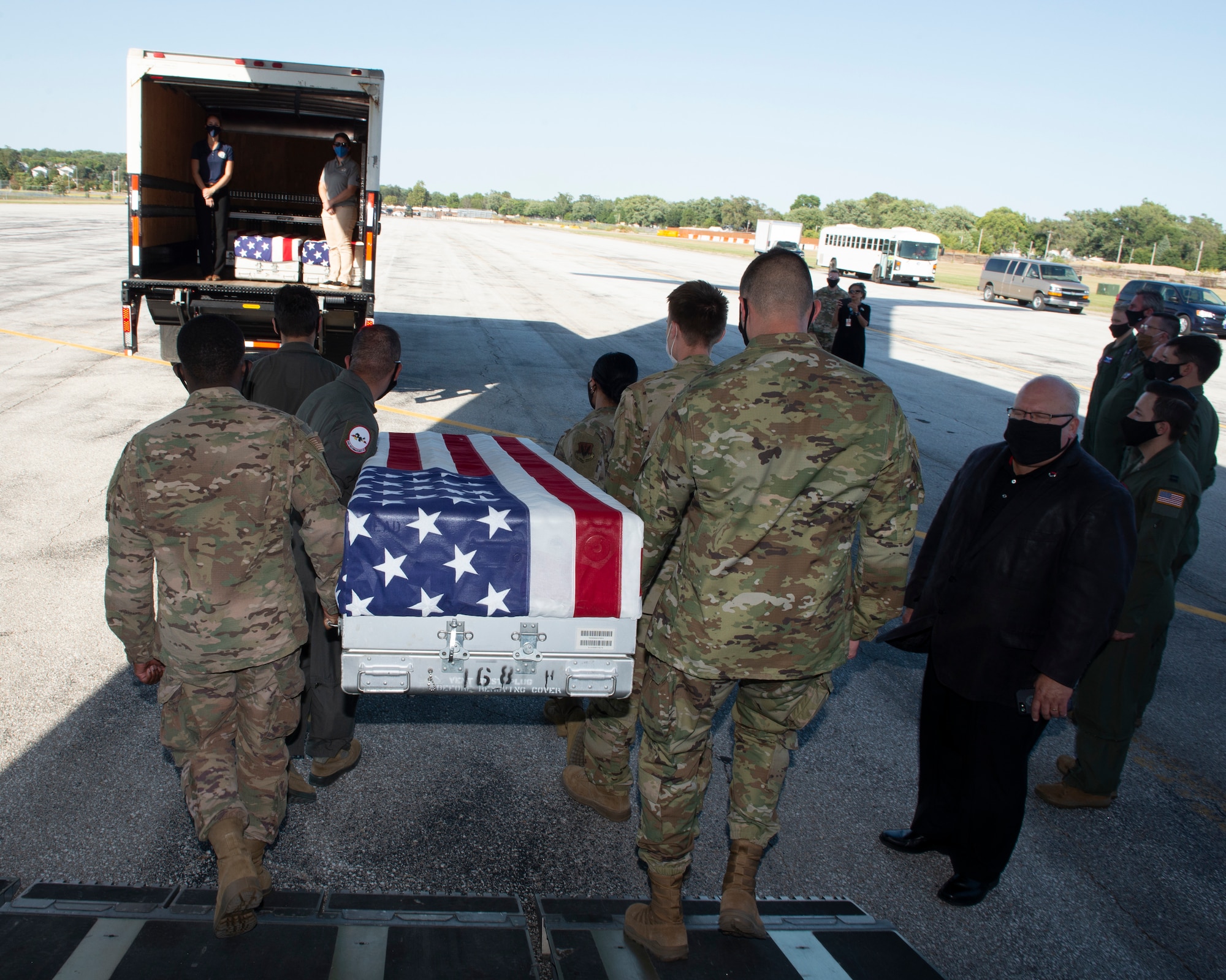 in foreground six Airmen walking off airplane ramp carrying a U.S. flag draped transfer case, walking to a transportation vehicle in the background