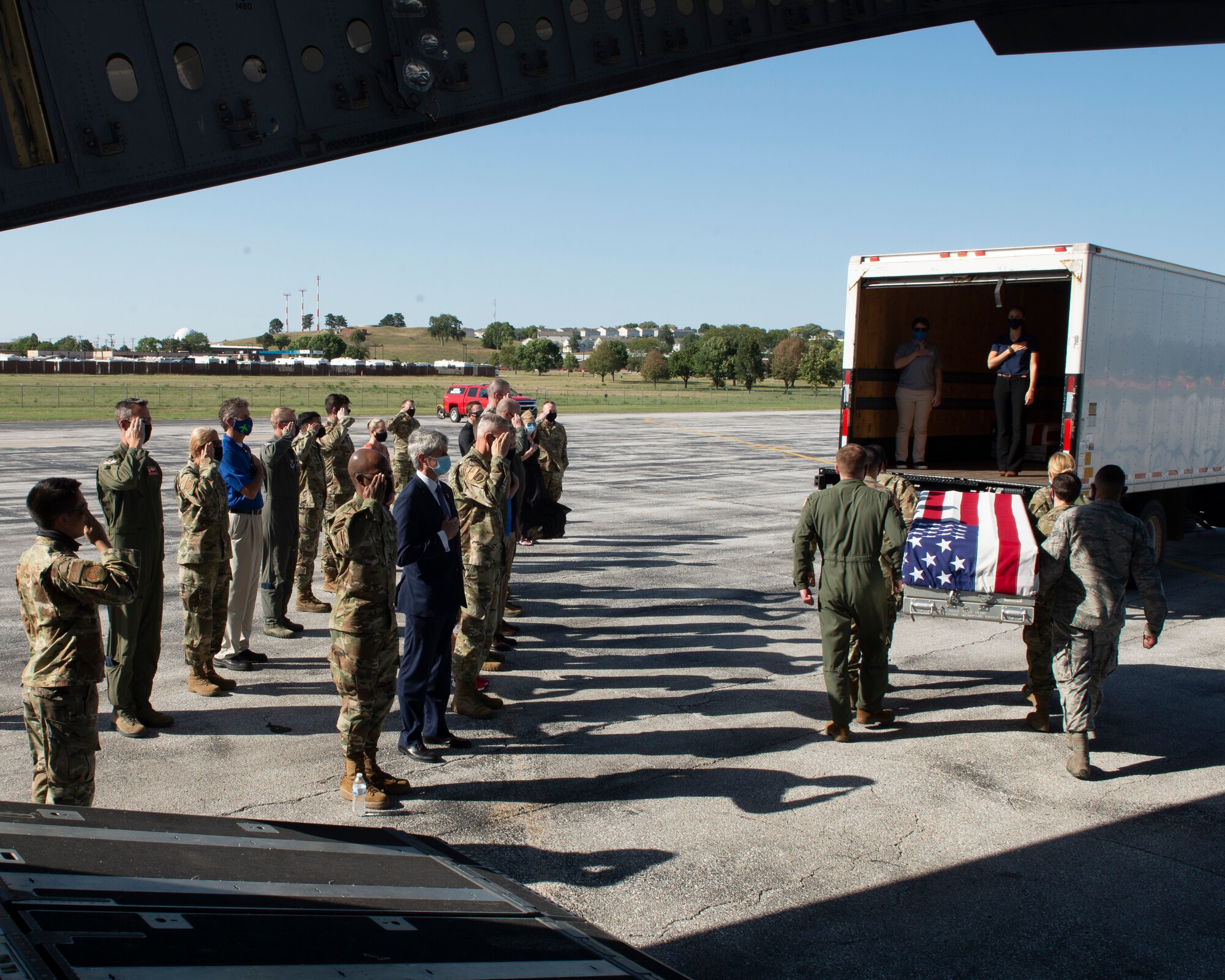 military members and civilians saluting as six Airmen carry a U.S. draped transfer case to a transportation vehicle