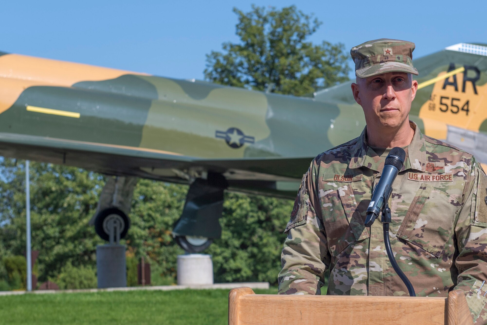 An Airman stands behind a podium in front of an aircraft static display.