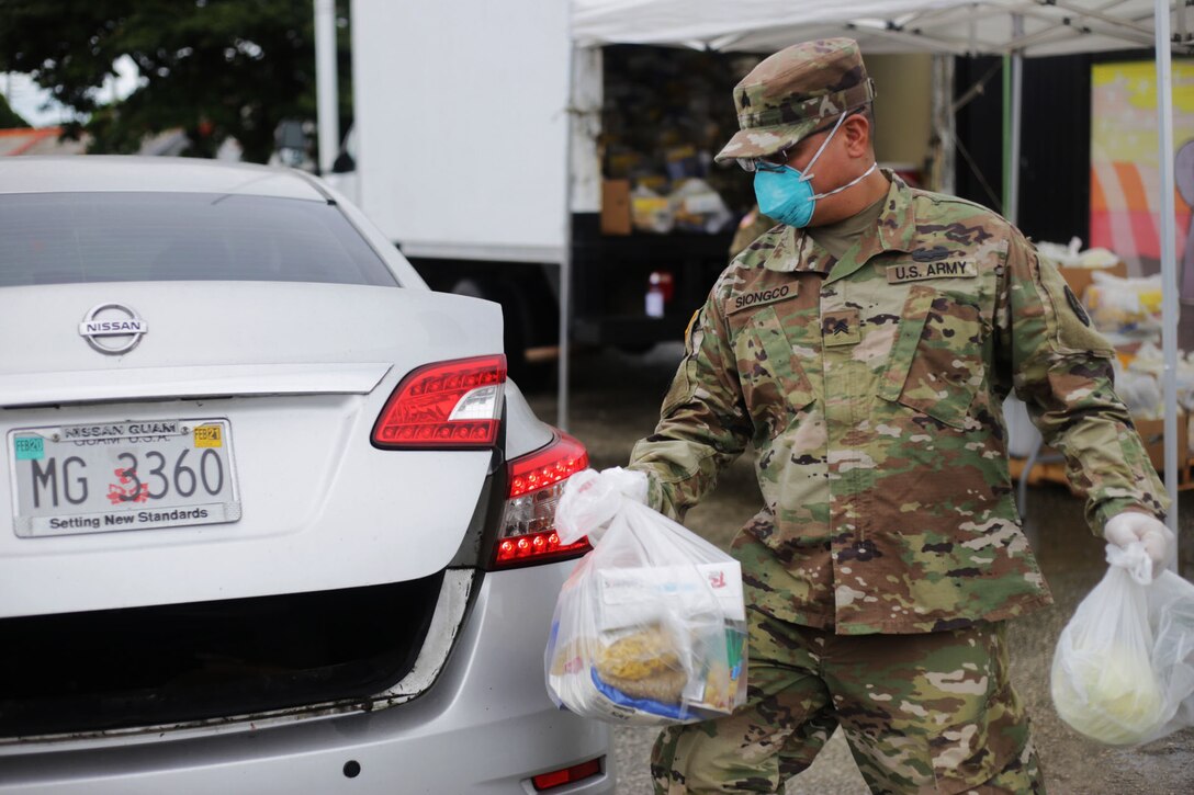 A soldier wearing a face mask carries bags of nonperishable goods, fresh fruits and vegetables to a car.