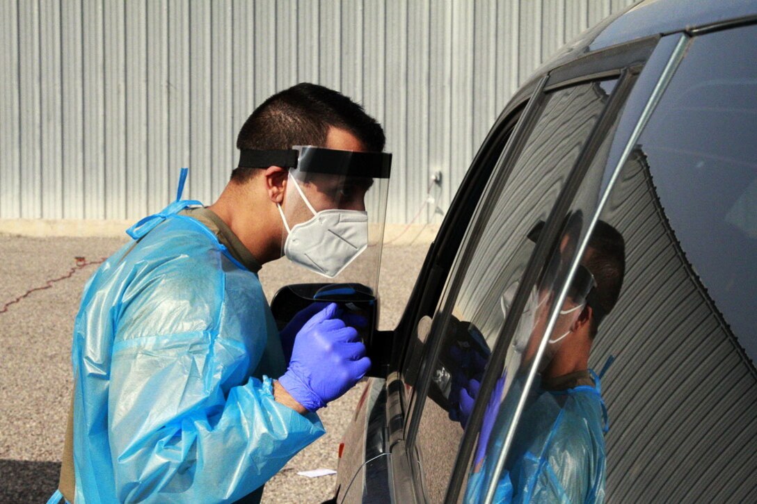 A man wearing personal protective equipment looks through the window of a car.