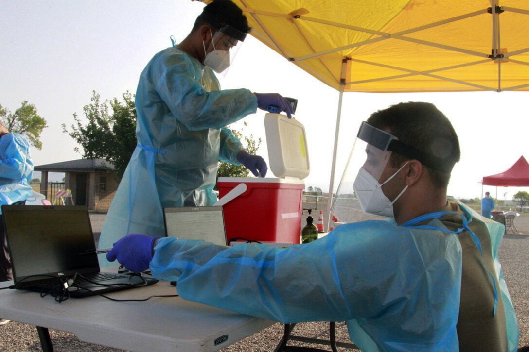 A man wearing personal protective equipment sits outside at a table and hooks up a laptop; another man stands at the table and holds open the lid of an ice chest.