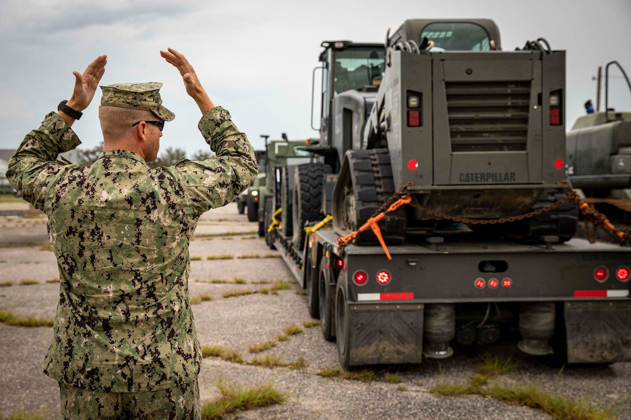 A sailor signals other sailors in military vehicles.