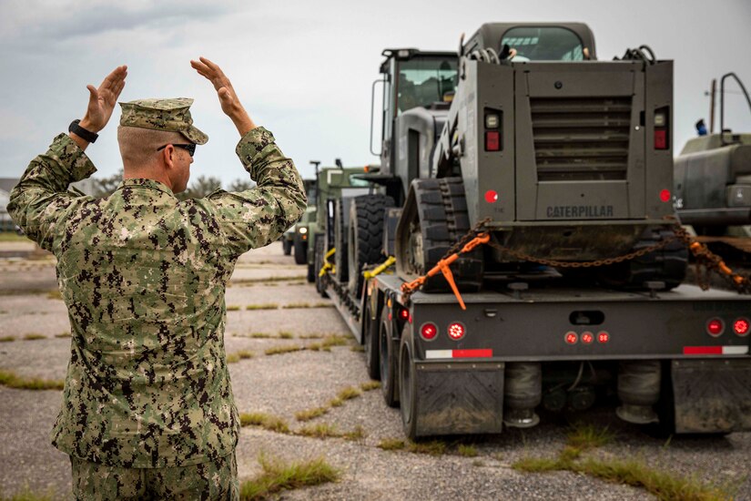 A sailor signals other sailors in military vehicles.