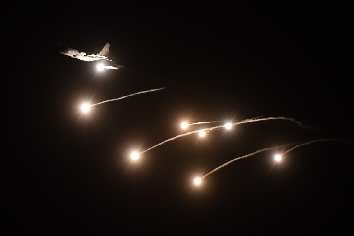 Flares shoot through a dark sky as a military aircraft flies upwards .