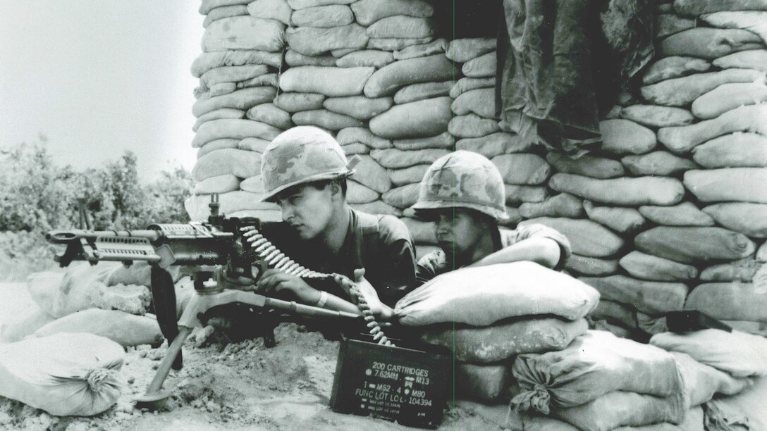 Black and white image of two male soldiers, one peering through the scope of a machine gun, surrounded by sandbags