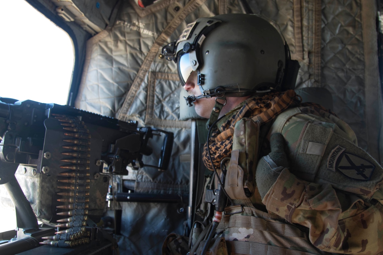 A soldier looks out the window of a helicopter. He sits near a machine gun.