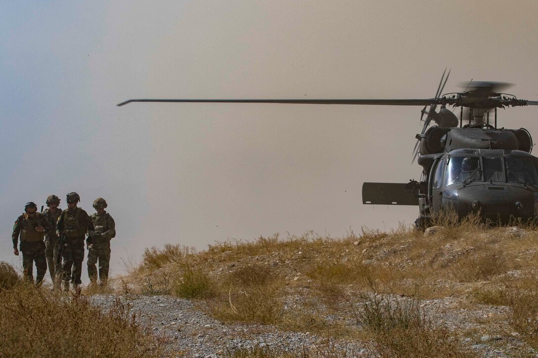 Soldiers walk down a gravel path. There is a helicopter nearby.