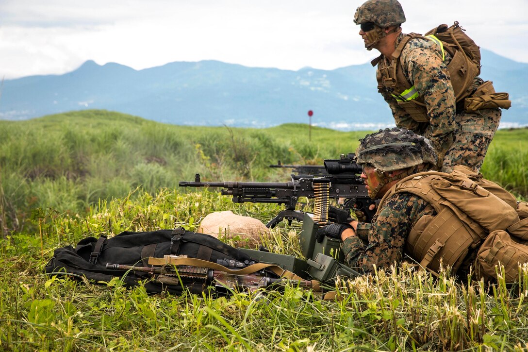 Marines holds a weapon and another Marine watches with mountains in the background.